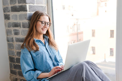 Happy casual beautiful woman in glasses working on laptop sitting on windowsill