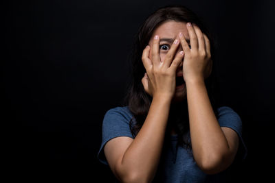 Portrait of scared young woman hiding face standing against black background