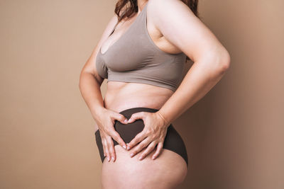 Self loving woman making heart shape on thigh on beige background