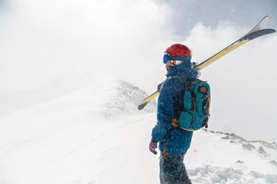 Man stands with his back to the camera with alpine skis on his shoulder against the backdrop of a