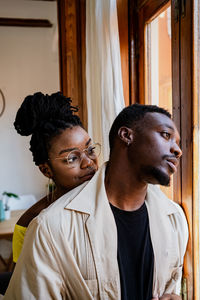 Cheerful young african american female in transparent eyeglasses with bunch looking at thoughtful man standing near window