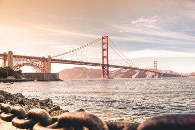 Suspension bridge over sea against cloudy sky during sunset