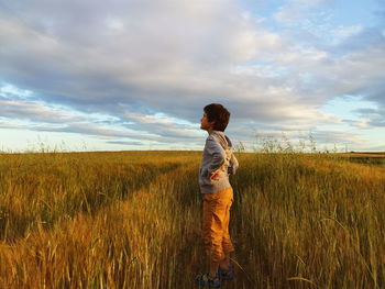 Side view full length of boy standing on grassy field against cloudy sky