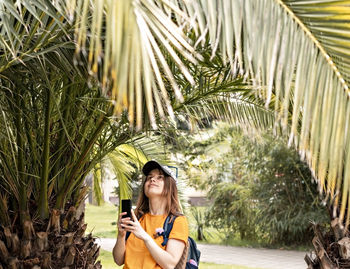 Young smiling woman tourist in cap taking photo on smartphone of tropical plant palm tree cicas 