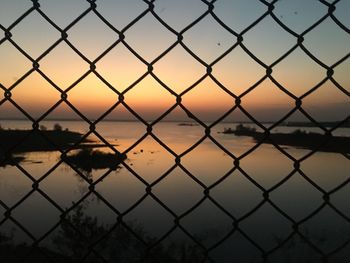 Silhouette chainlink fence by river against sky during sunset