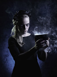 Portrait of spooky young woman holding bowl emitting smoke against black background