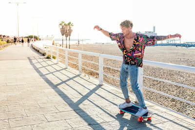 Stylish male hipster in trendy wear riding modern skateboard with outstretched arms while balancing and enjoying weekend in summer