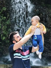 Father picking up son while standing against waterfall