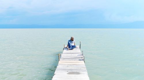 Mature man sitting on pier over sea against sky during sunny day