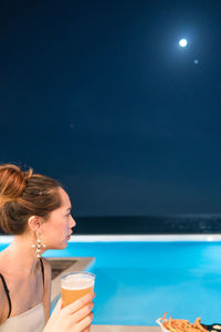 Young woman holding beer glass by swimming pool at restaurant against sky