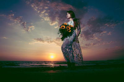 Full length of woman with flowers standing on sea shore against sky