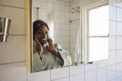 Happy mature woman touching face while looking herself in bathroom mirror at home