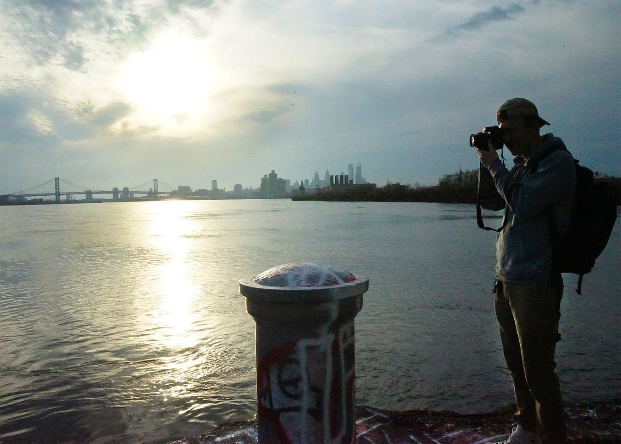 MAN PHOTOGRAPHING BY RIVER IN CITY AGAINST SKY DURING SUNSET
