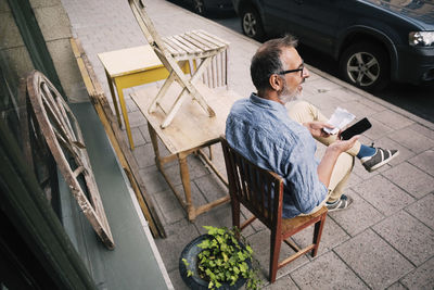 High angle view of man holding mobile phone while sitting outside antique shop