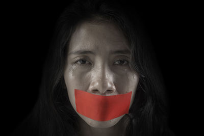 Portrait of crying woman mouth sealed with red tape against black background