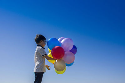 Boy holding multi colored balloons while standing against clear blue sky