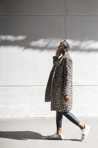 Side view of african american female in coat talking on cellphone while standing near building on sunny street in city