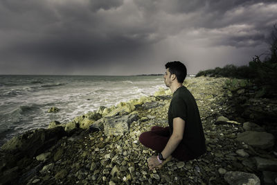 Man sitting on rocks at beach against sky