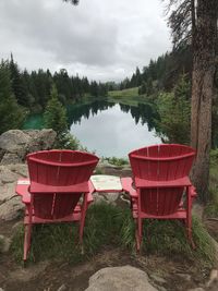 Red chairs by trees on lake against sky