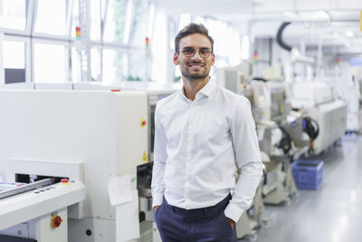 Smiling young businessman standing with hands in pockets at illuminated factory