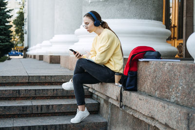 Portrait of casual student girl in headphones using mobile phone app outdoors. young woman student