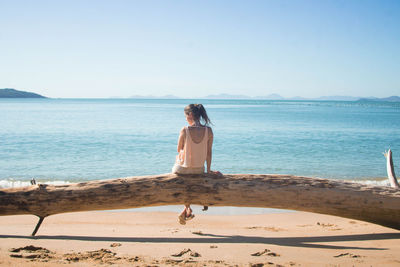 Rear view of woman sitting on fallen tree at beach against clear sky
