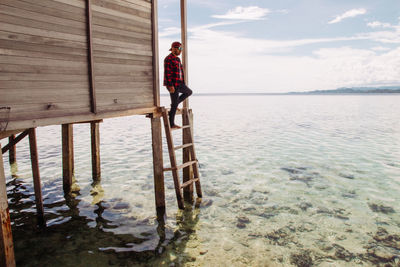 Man moving down on ladder of stilt house in sea against sky