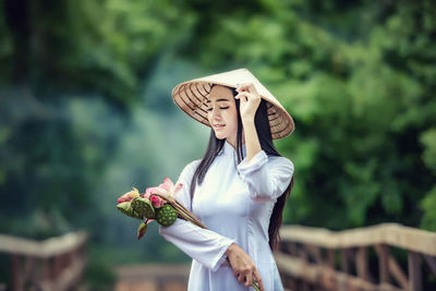 Woman wearing conical hat while holding flowers and pods against trees