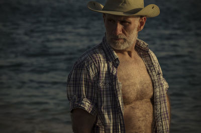 Adult man in cowboy hat with open shirt on beach against sea