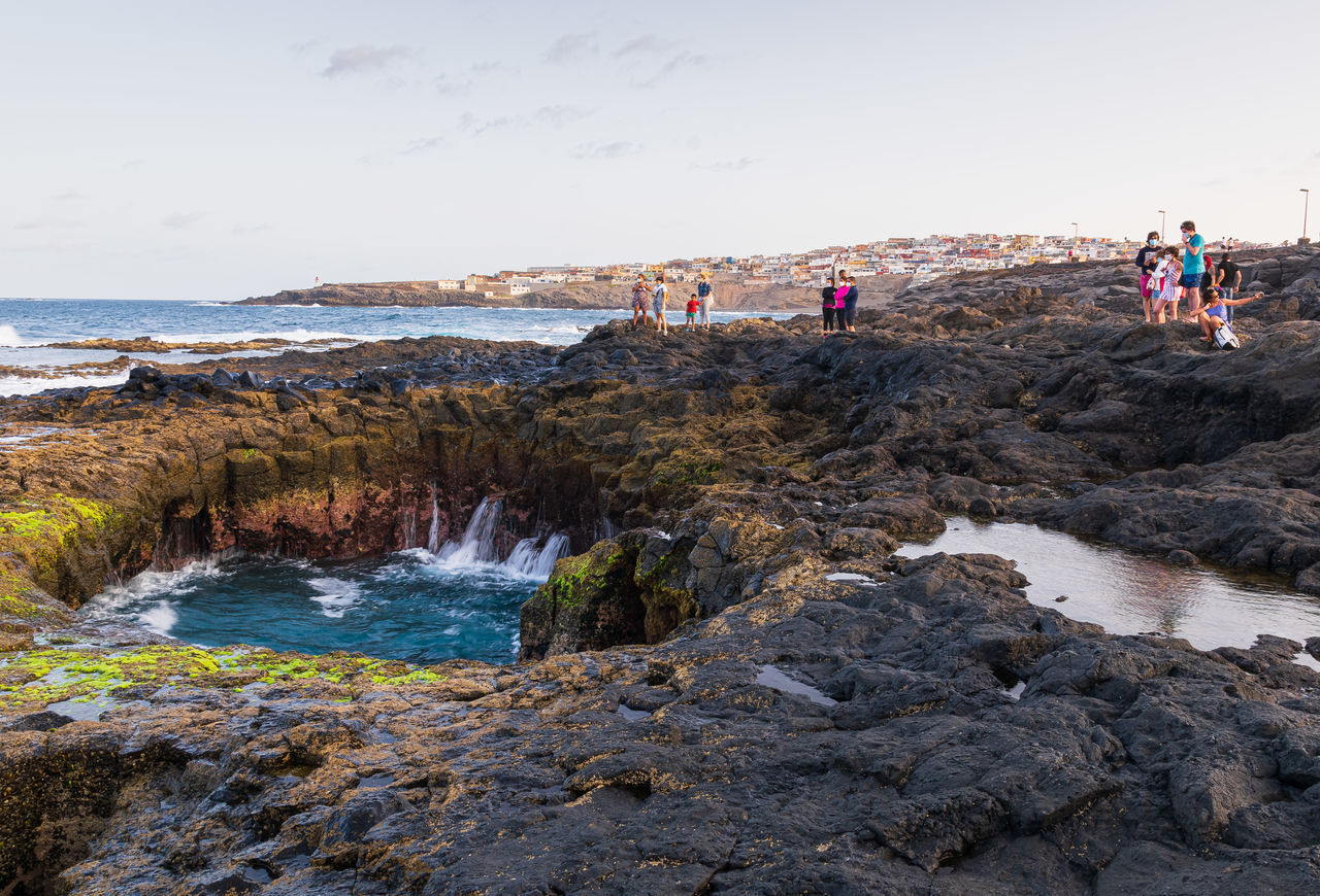 GROUP OF PEOPLE ON ROCKS BY SEA AGAINST SKY