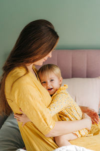 A young woman in a yellow dress sitting on the bed gently hugs her child. 