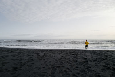 Man in a yellow jacket standing on the black sand beach overlooking the horizon
