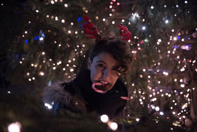 Happy young woman eating candy cane by illuminated christmas tree at night