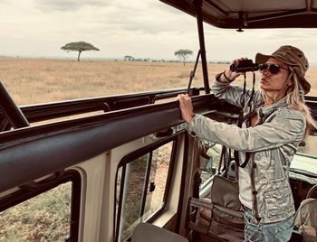 Woman holding binoculars while standing in jeep against sky