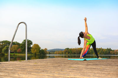 Mature woman exercising on pier over river against sky