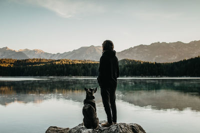 Rear view of man with dog looking at lake against mountain range