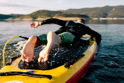 Back view of unrecognizable barefoot male in wetsuit lying on paddle board and swimming on lake surface while practicing water sport in summer day