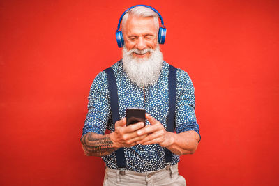 Happy bearded man using mobile phone while wearing headphones against red background
