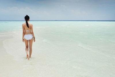 Rear view of young woman in bikini standing at beach against sky