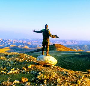 Rear view of young man with arms outstretched standing on mountain against sky during sunset