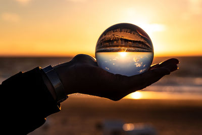 Close-up of hand holding crystal ball against sea during sunset