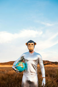 Anonymous guy in silver latex suit with geometric monkey mask looking at camera and holding globe in nature