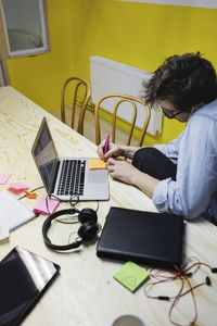 High angle view of young man using smart phone while writing on adhesive note at desk in board room