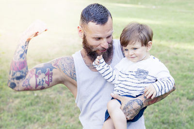 Smiling bearded man flexing muscle while holding son on field 