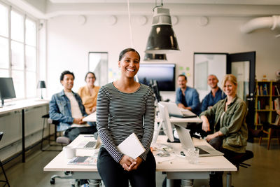 Smiling businesswoman sitting at desk while colleagues in background