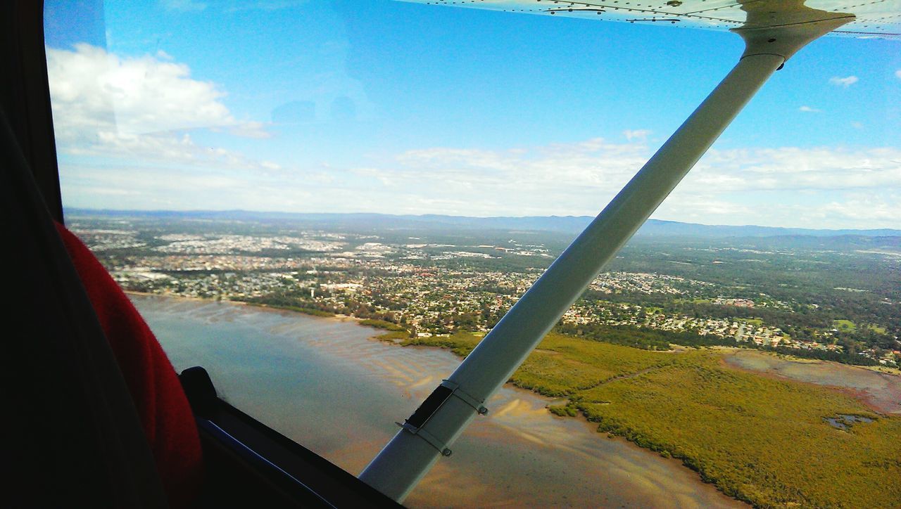 airplane, transportation, mode of transport, flying, air vehicle, window, sky, city, vehicle interior, cityscape, travel, part of, on the move, aircraft wing, mid-air, journey, cloud - sky, aerial view, cropped, transparent, water, crowded, river, cloud, day, blue, wide shot, sea, outdoors, horizon over land, nature