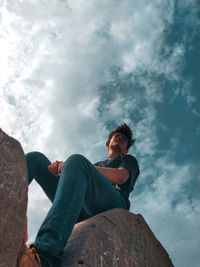 Low angle view of young man sitting on rock against sky