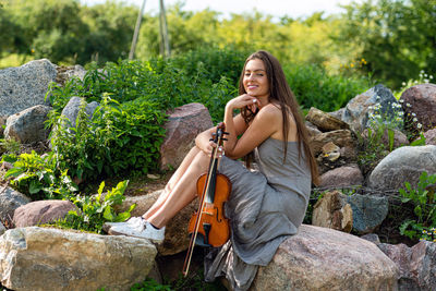 Beautiful brunette in a linen dress with a violin on a pile of boulders at an old country house