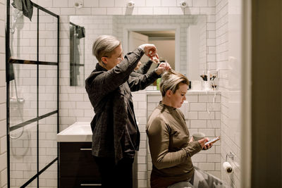 Side view of lesbian woman cutting hair of girlfriend in bathroom at home