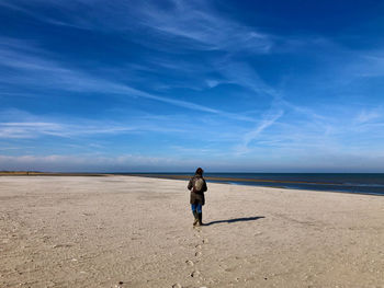 Rear view of a woman walking on an empty beach under a blue sky with white cloud streaks